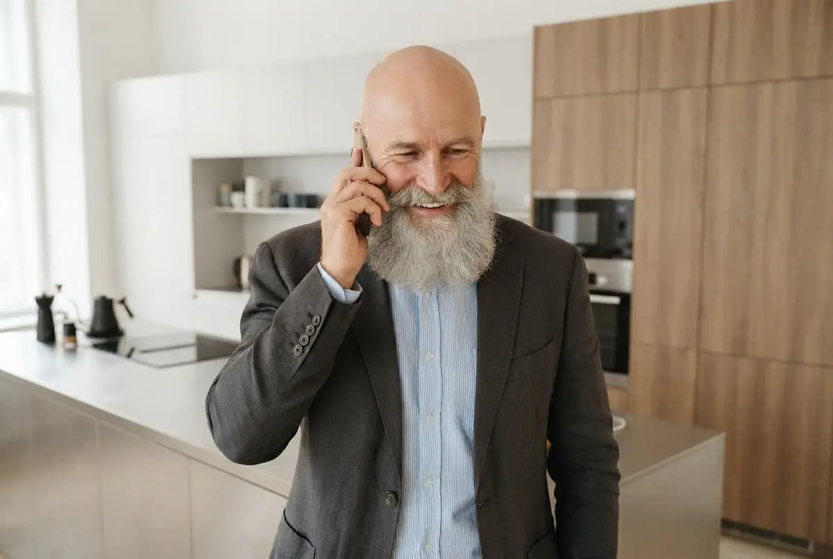 Hombre con barba hablando por teléfono y sonriendo en su cocina