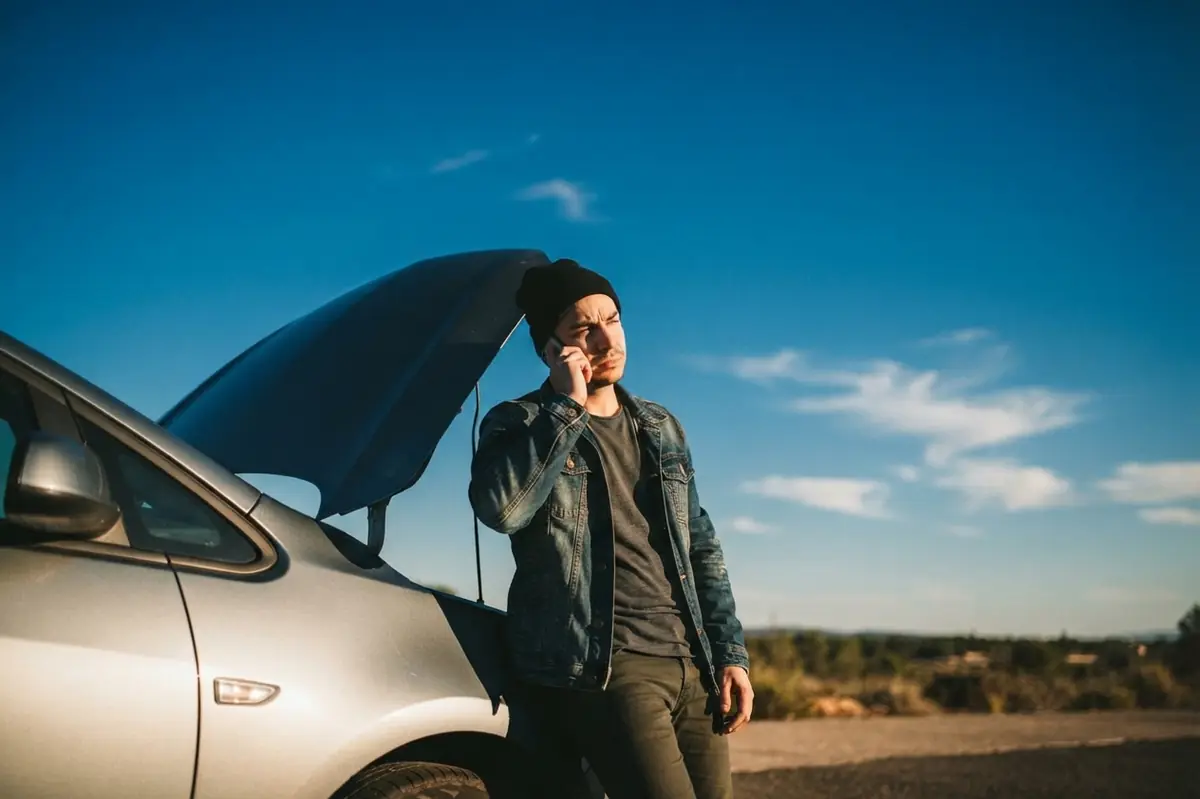 Conductor llamando por teléfono junto a su coche averiado con el capó abierto