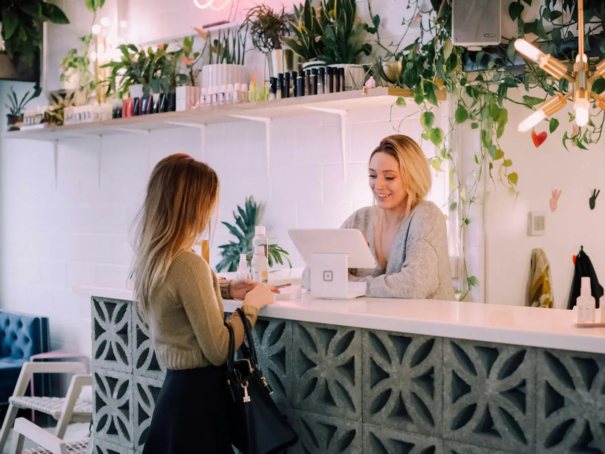 Recepcionista sonriendo detrás del mostrador de un salón lleno de plantas mientras atiende a una clienta