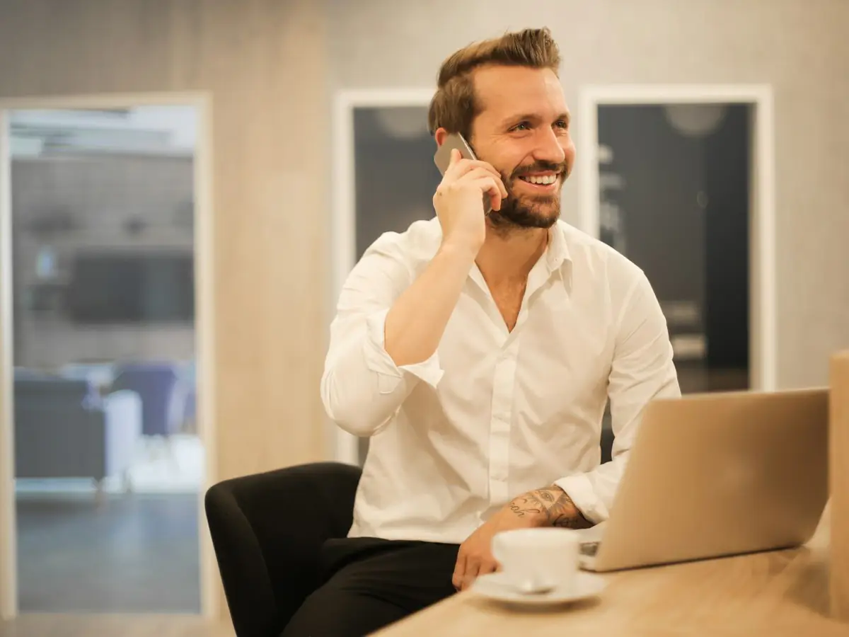 Hombre sonriendo al teléfono con el portátil y un café delante