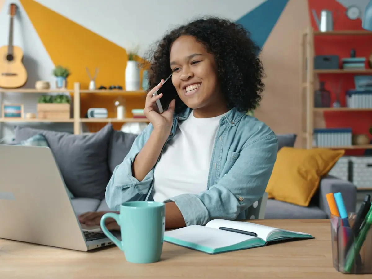 Chica joven sonriendo al teléfono delante del portátil, en un salón luminoso